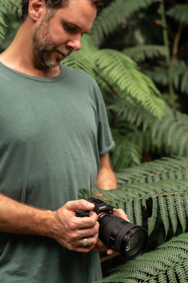 David Lloyd looking down while holding a Sony camera, surrounded by tropical foliage | About Page Portrait