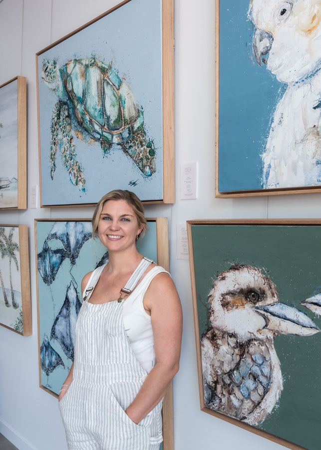 Stephanie Elizabeth – standing against gallery wall with her original paintings displayed behind her | About SEA Gallery