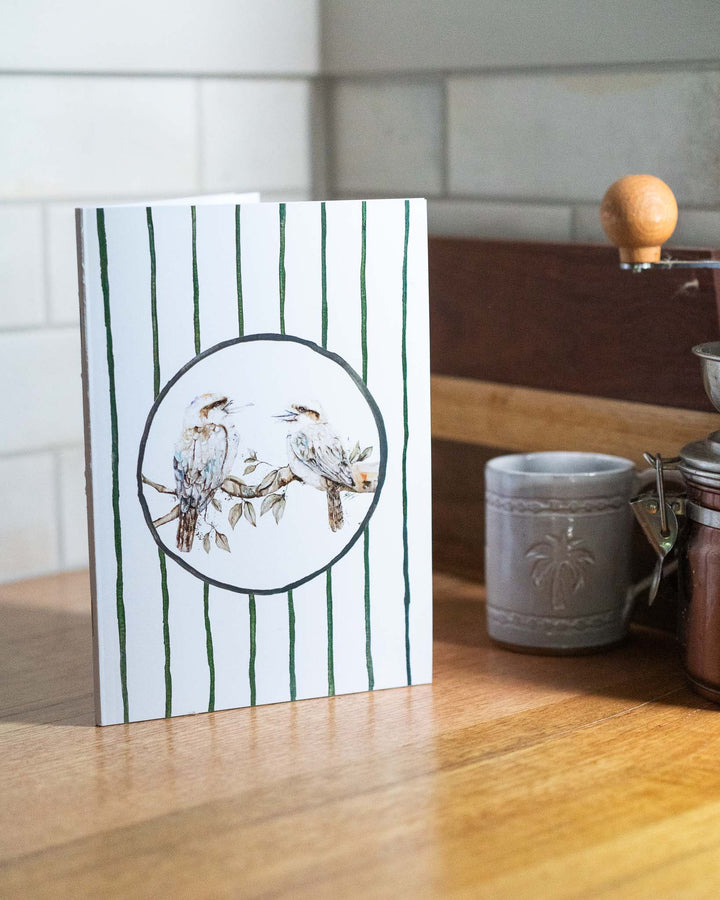 “Branching Out Together” A5 notebook with kookaburras on a wooden counter next to a mug.