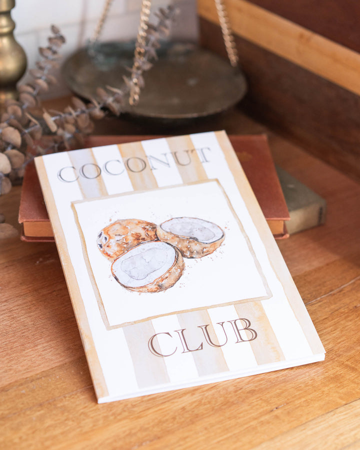 Coconut Club A5 Lined Notebook with three coconuts resting on a wooden surface.