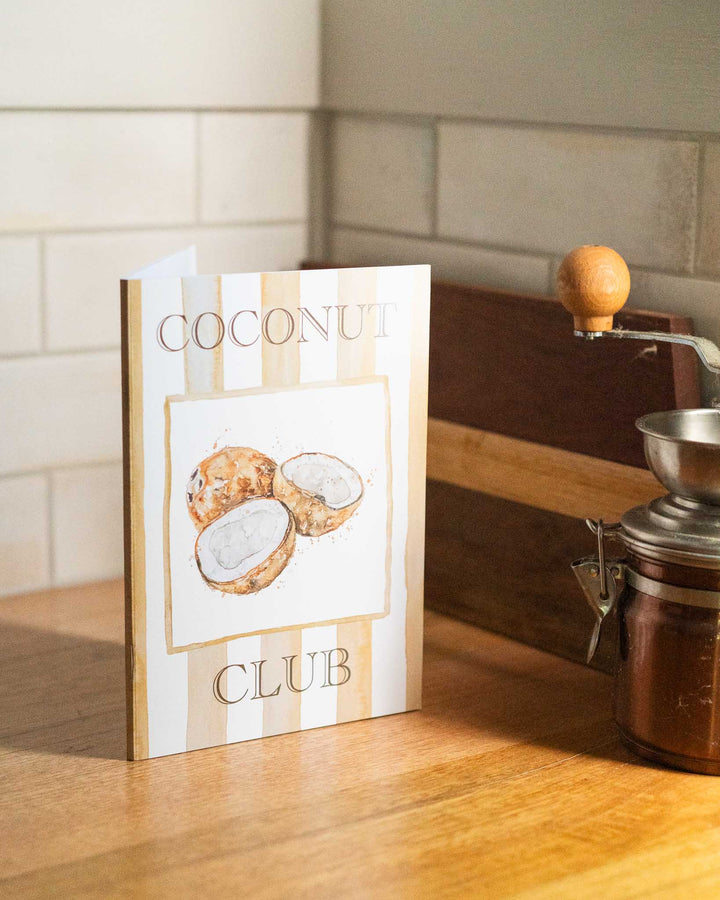The Coconut Club A5 Lined Notebook rests on a sunlit counter with tropical decor and bright natural light.
