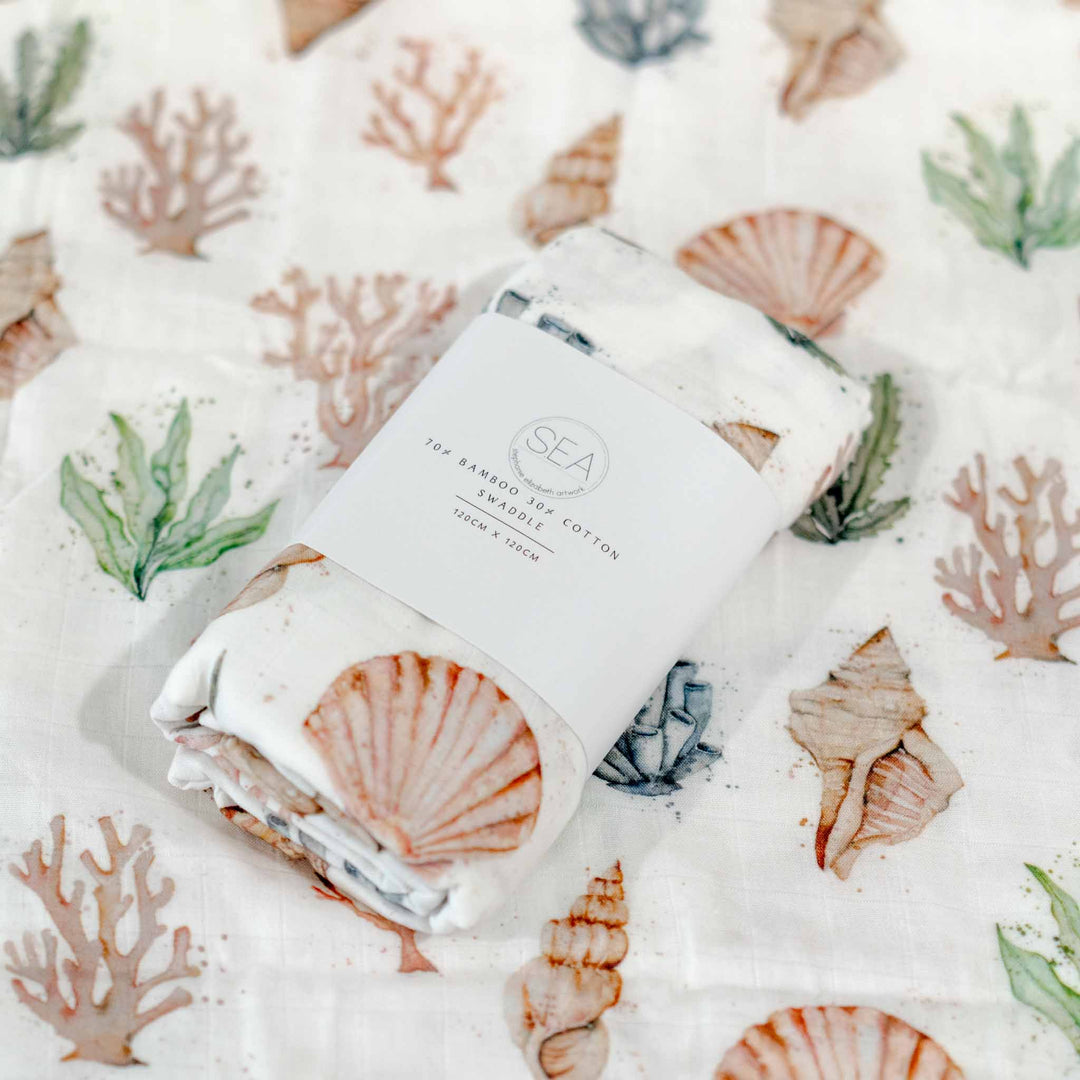 A folded bamboo- cotton muslin cloth with a watercolour seashell and coral print, wrapped in a white paper band labeled &