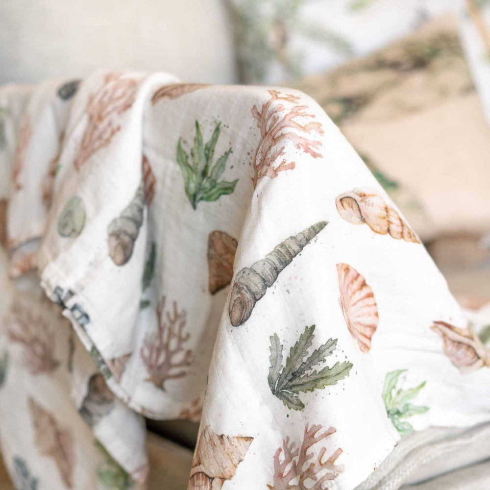 A close-up of our draped coral reef  white muslin blanket featuring a watercolor print of seashells, coral, and green sea plants, styled on a sofa with a matching pillow in the background.