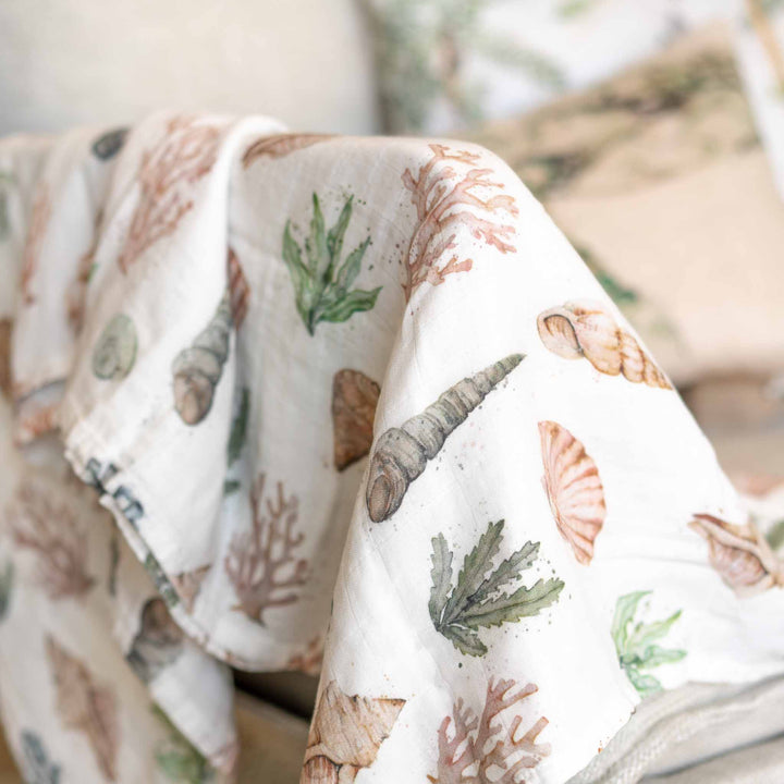 A close-up of our draped coral reef  white muslin blanket featuring a watercolor print of seashells, coral, and green sea plants, styled on a sofa with a matching pillow in the background.