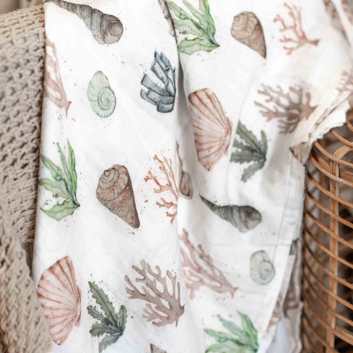 Our white bamboo/ cotton muslin baby blanket with a watercolor print of seashells, coral, and seaweed, draped over a woven wicker basket. Inspired by hte Great Barrier Reef.