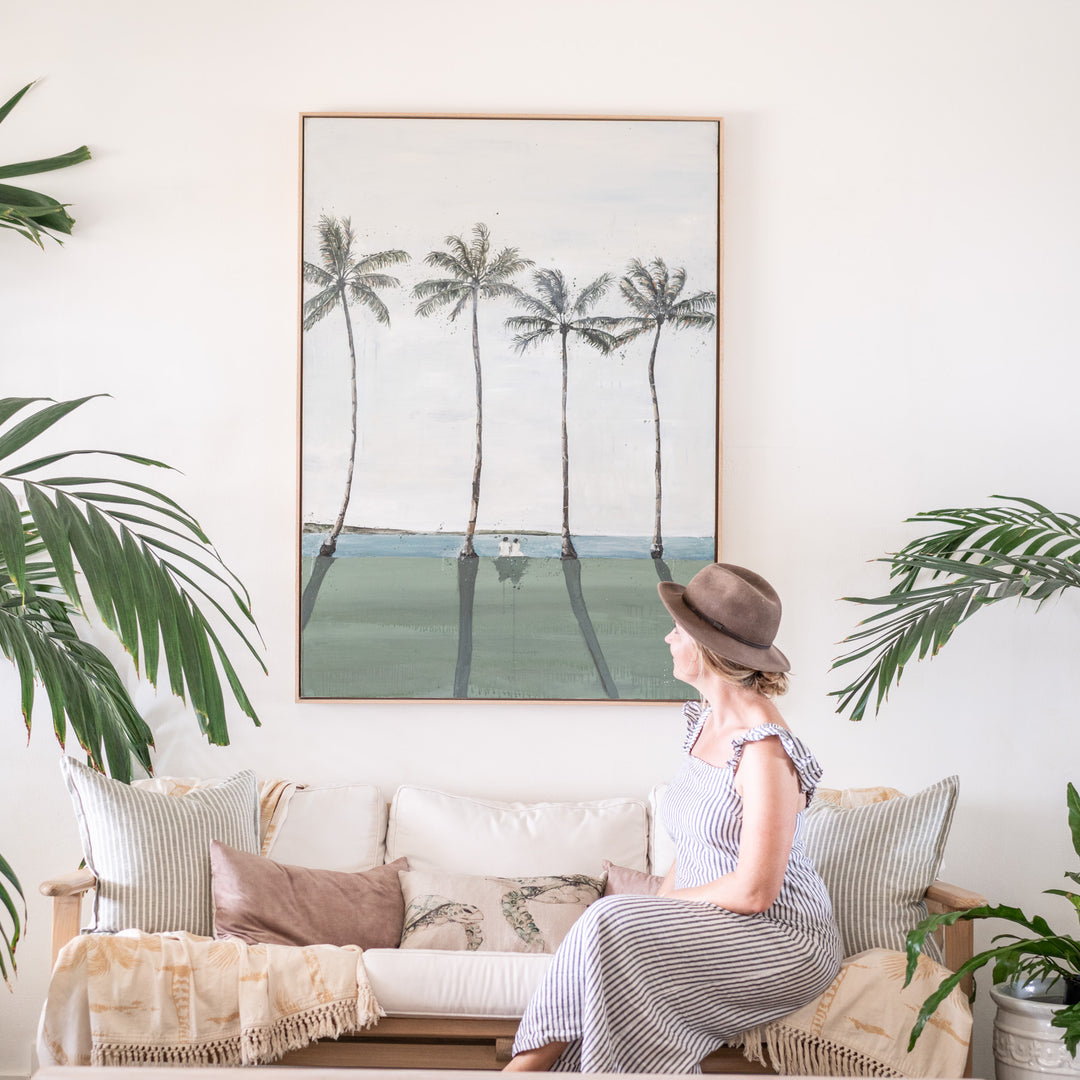 Just Us – original coastal artwork by Stephanie Elizabeth photographed in a tropical interior with the artist seated beneath the framed painting