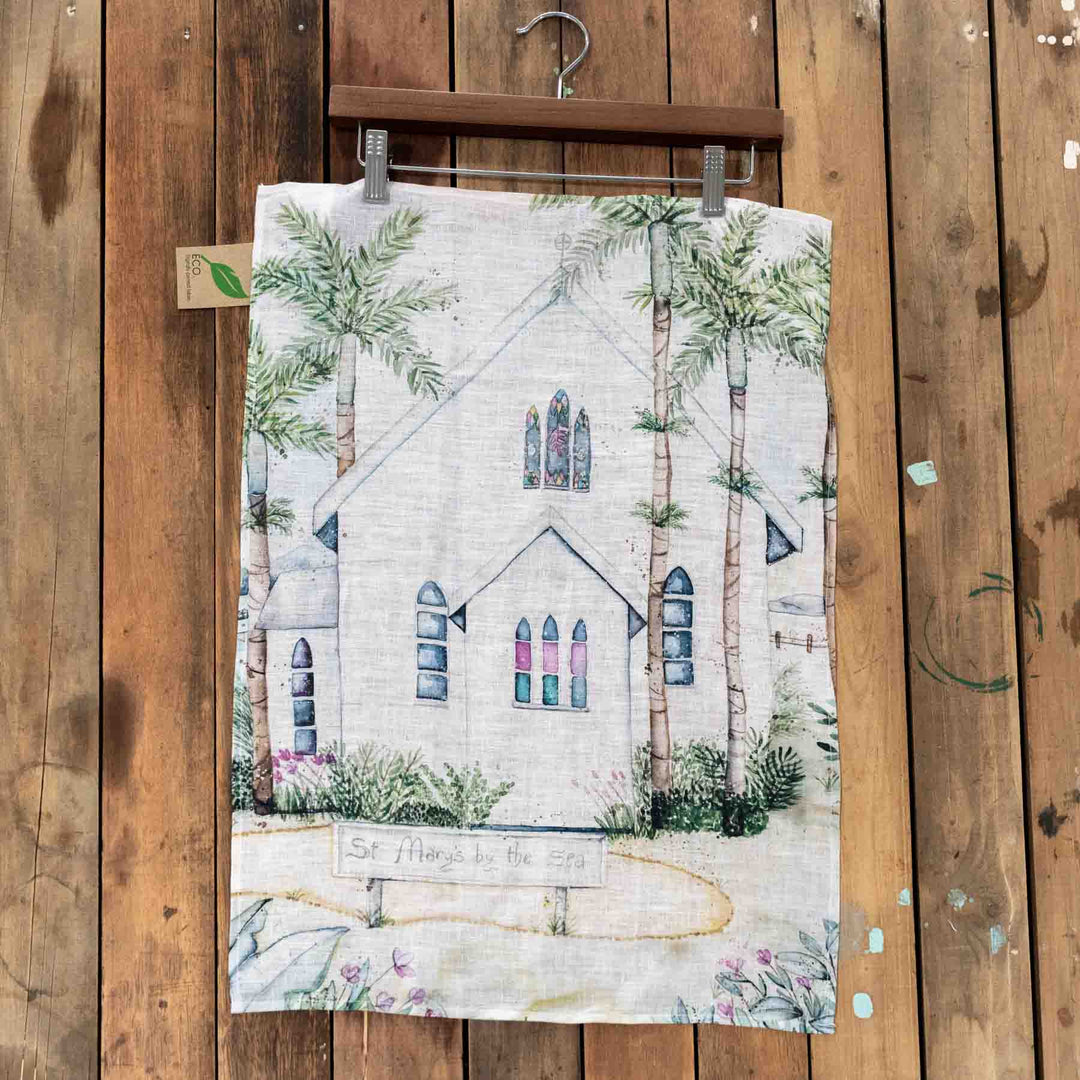 St Marys linen tea towel featuring a white church and palm trees, displayed on a rustic wooden wall.