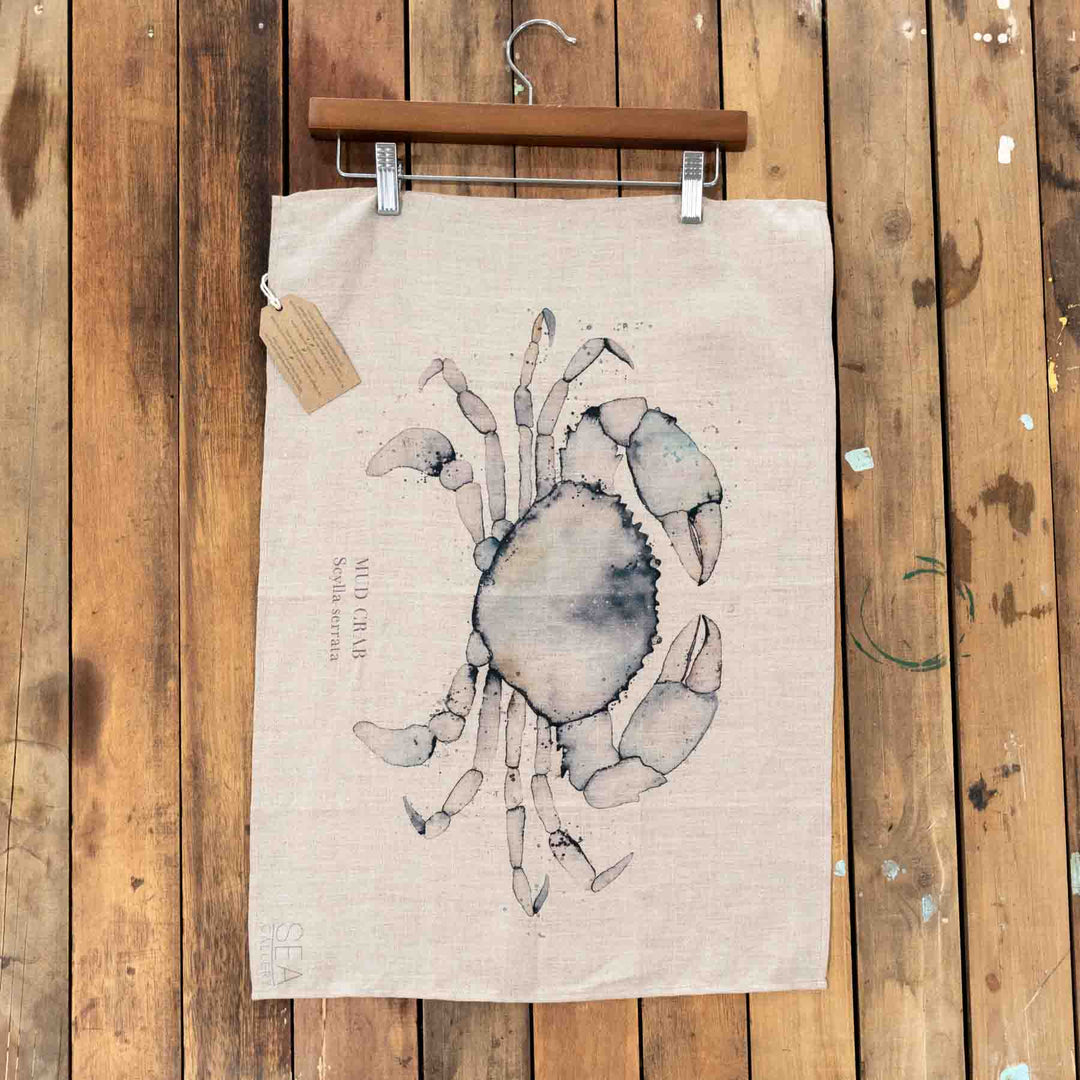 Mud Crab Linen Tea Towel hangs on a wooden hanger in front of a natural wood background.