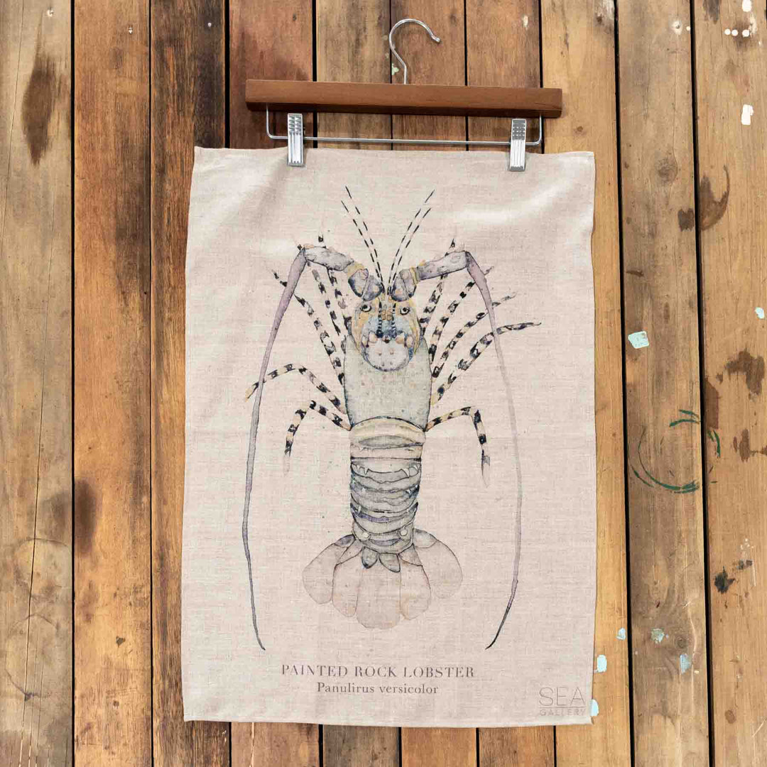 The Lobster linen tea towel with a red lobster design displayed against a wooden wall.