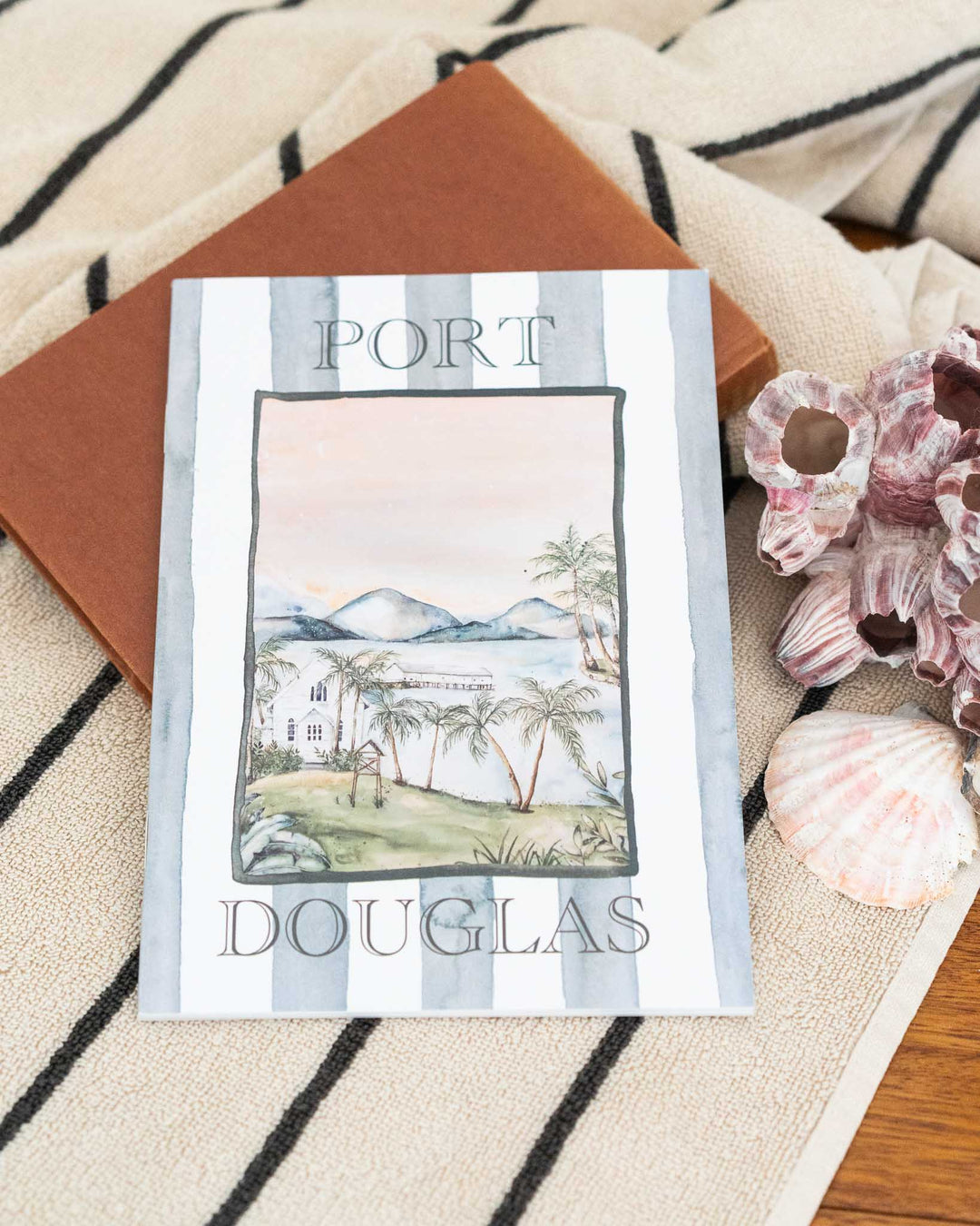 Port Douglas A5 lined notebook next to seashells and a brown book on a towel.