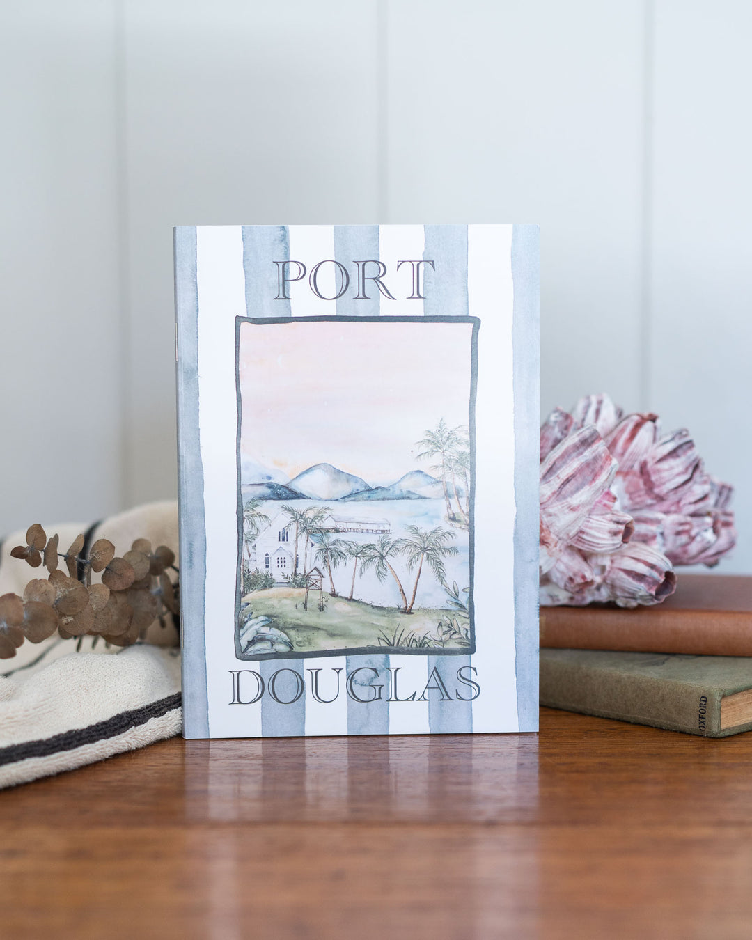 Port Douglas A5 Lined Notebook with coastal watercolor cover, placed beside books and flowers.