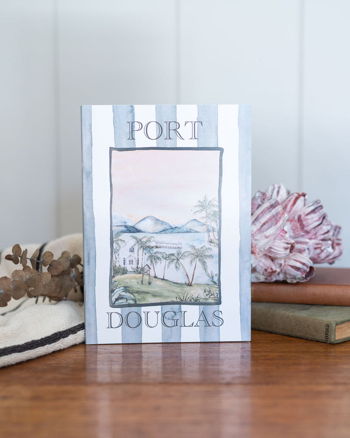 Port Douglas A5 Lined Notebook with coastal watercolor cover, placed beside books and flowers.
