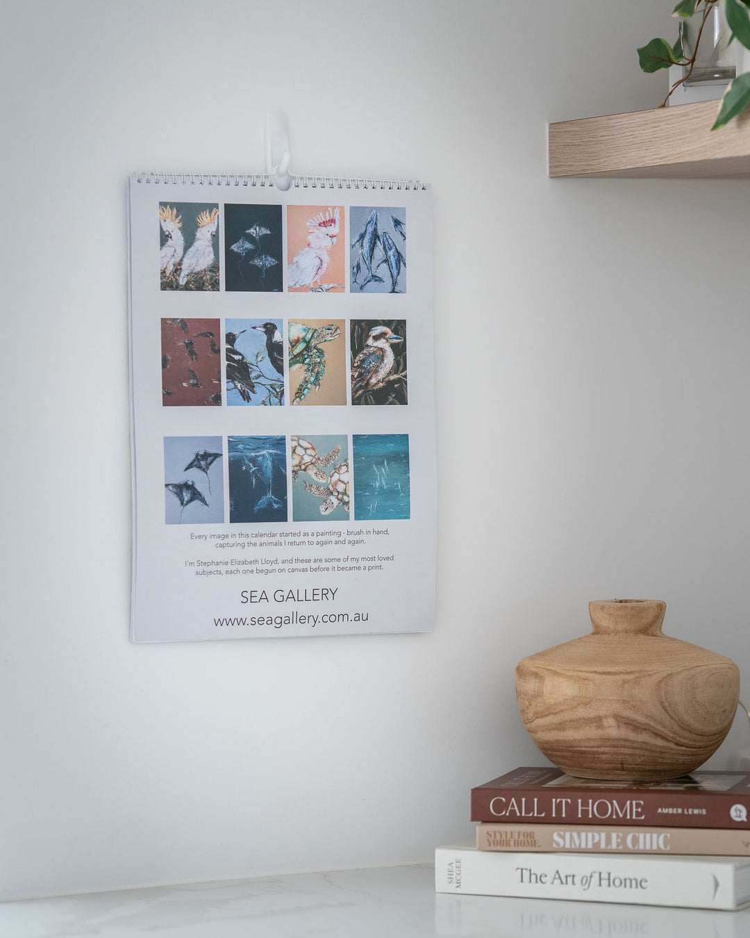 2027 Australian Wildlife Art calendar by Stephanie Elizabeth displayed above books and a vase on a white shelf.
