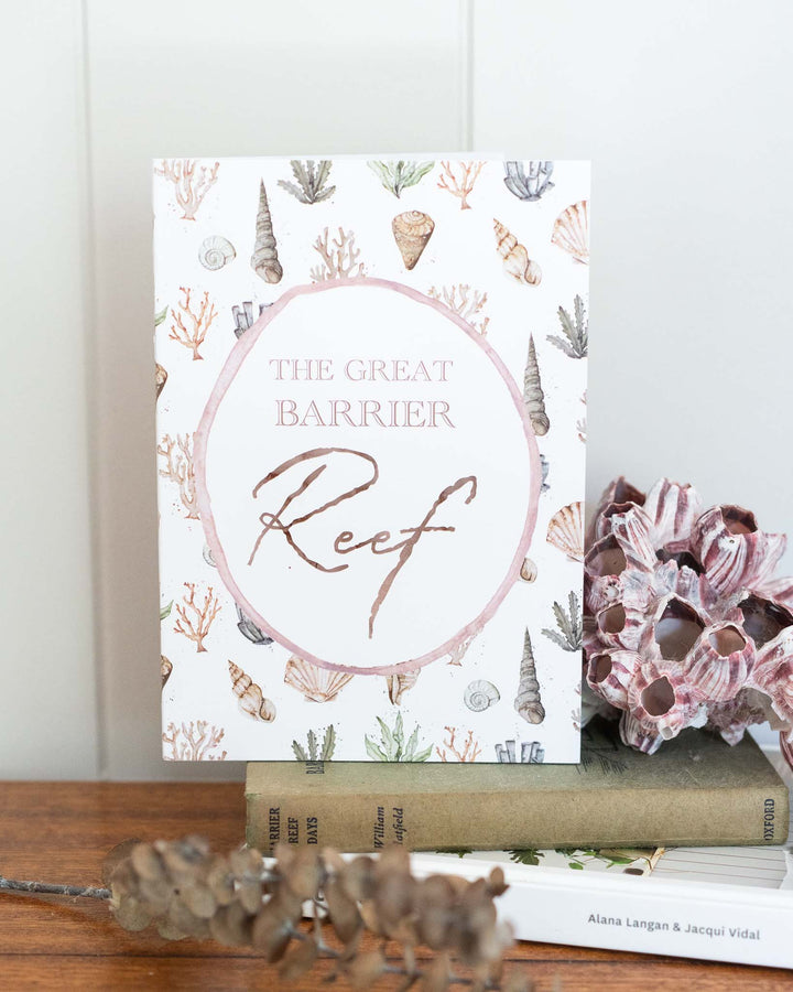 The Great Barrier Reef A5 lined notebook with seashell and coral artwork, resting on stacked books and wood.