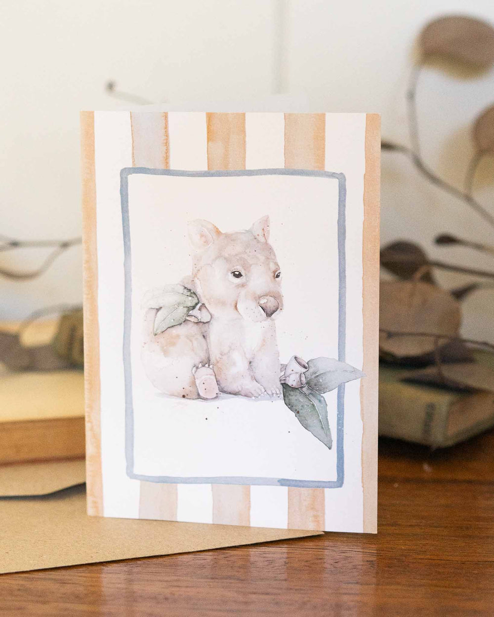 Wombat Woddle greeting card featuring a watercolor wombat with green leaves, displayed on a wooden table.