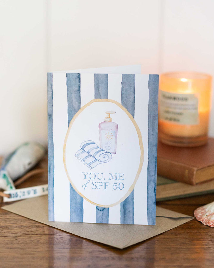 “You, Me and SPF 50” greeting card with sunscreen and towels on a colorful striped watercolor background.