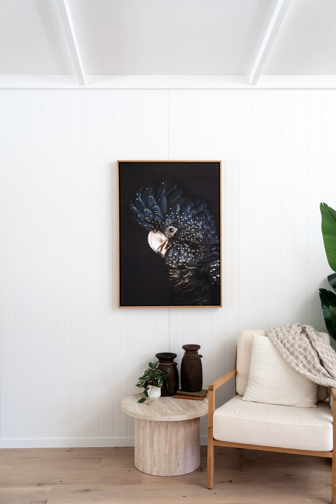 “A Little Less Conversation” black cockatoo artwork above an armchair, side table, and plants in a cozy room setting.