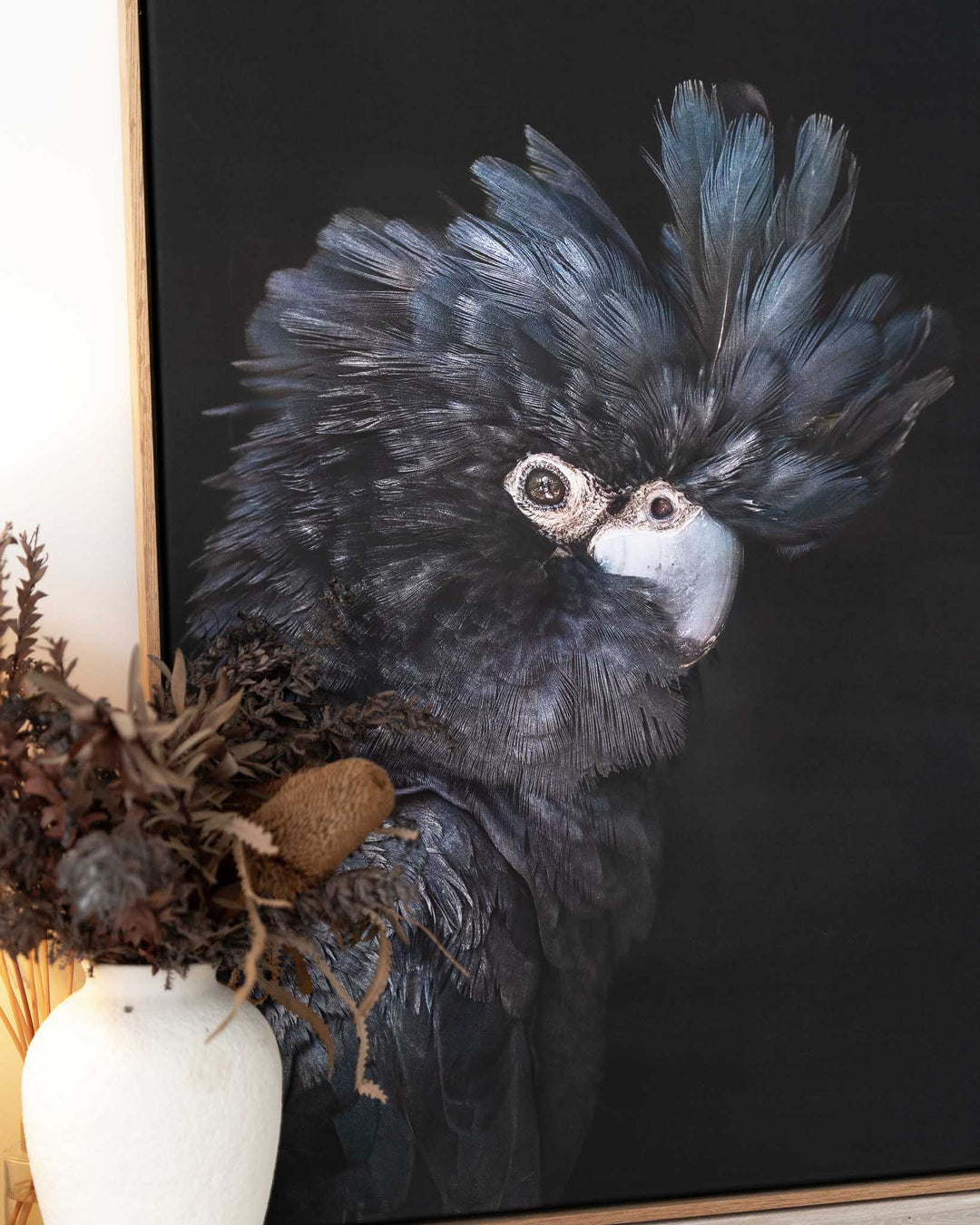 A Little More Action framed print featuring a black cockatoo, displayed next to a white vase with dried flowers.