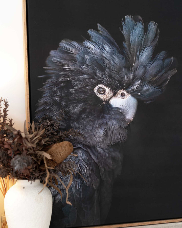 A Little More Action framed print featuring a black cockatoo, displayed next to a white vase with dried flowers.