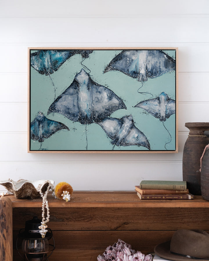 A Sea of Spots eagle rays fine art print by Stephanie Elizabeth, framed canvas in an oak frame above a bookshelf