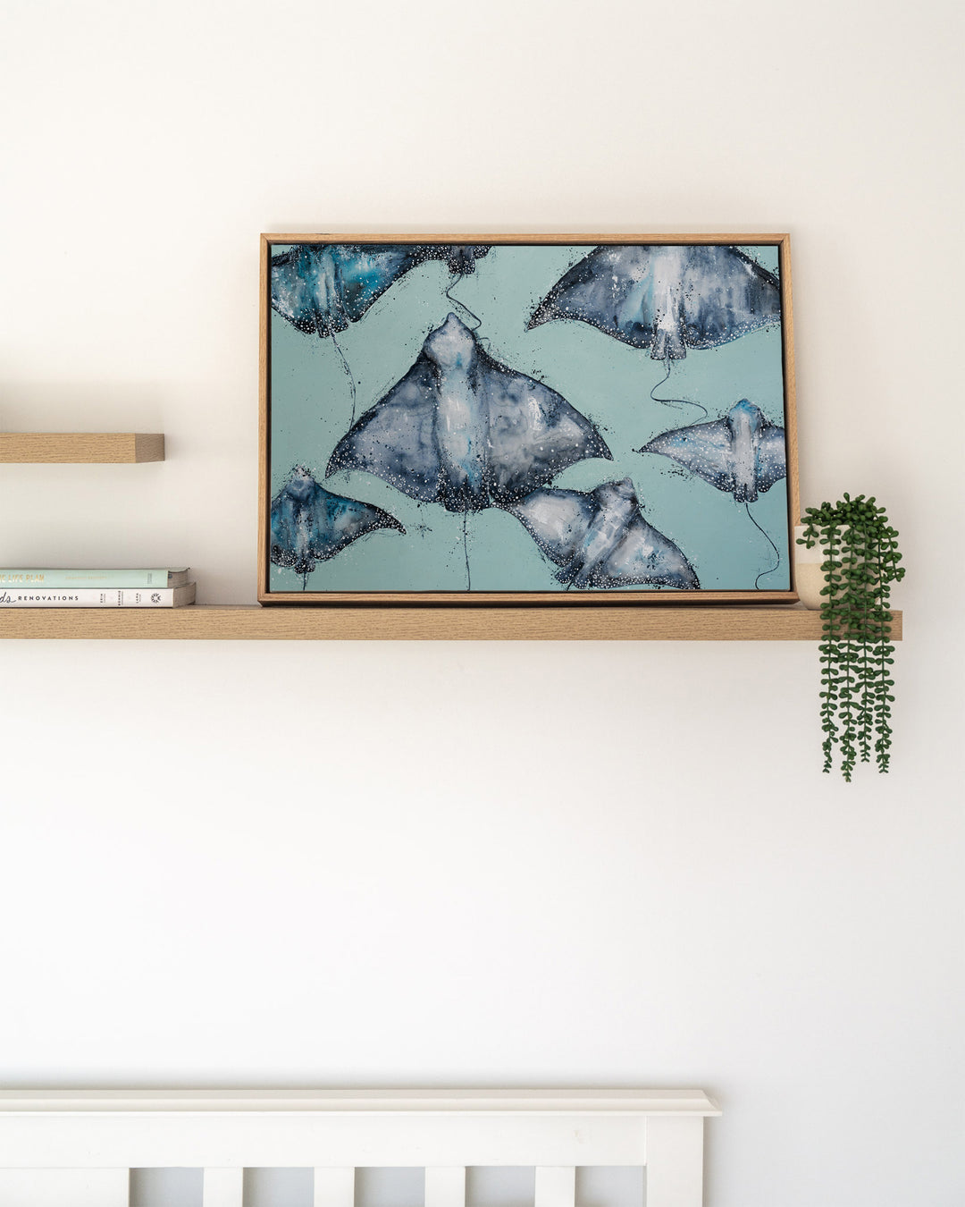 A Sea of Spots eagle rays fine art print by Stephanie Elizabeth, framed canvas in an oak frame on a shelf