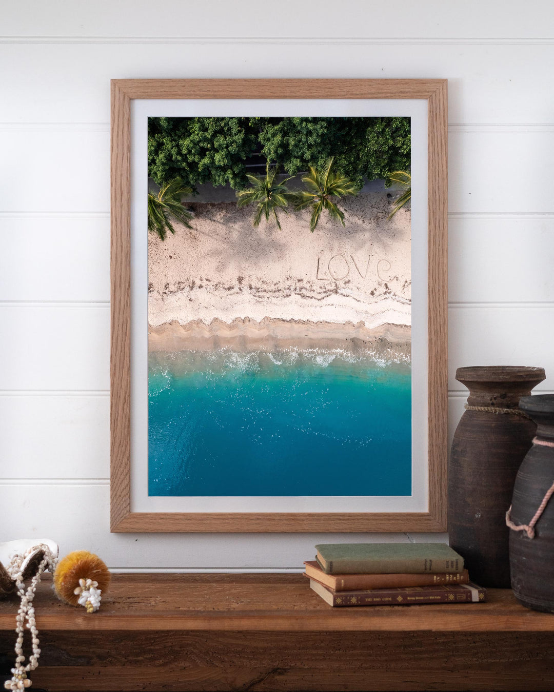 All You Need Is... Whitsundays aerial print of LOVE in the sand, framed on a white wall above a wooden shelf.