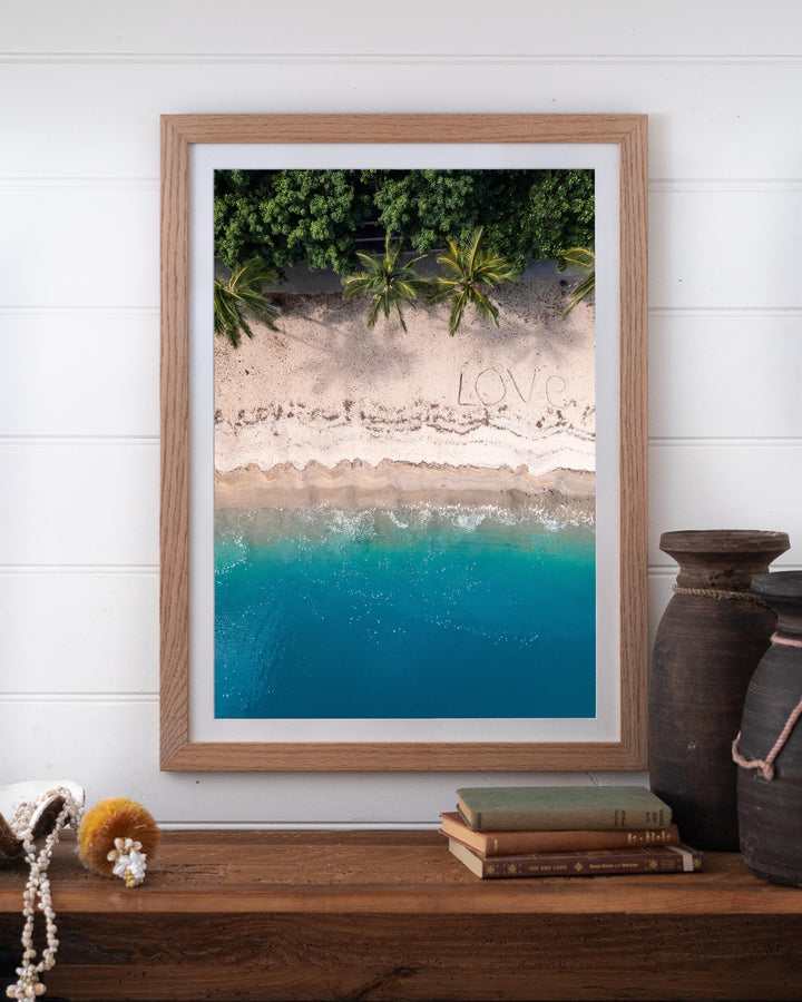 All You Need Is... Whitsundays aerial print of LOVE in the sand, framed on a white wall above a wooden shelf.