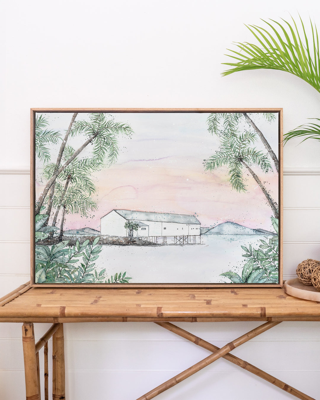 Balmy Beachside Sugar Wharf Port Douglas fine art print by Stephanie Elizabeth, framed canvas in an oak frame above a rattan table