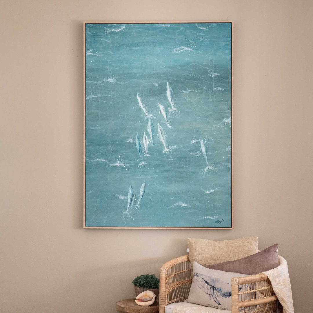 Bold and the Beautiful – coastal dolphin artwork in soft blue tones displayed above rattan chair in neutral interior setting | Original Artwork