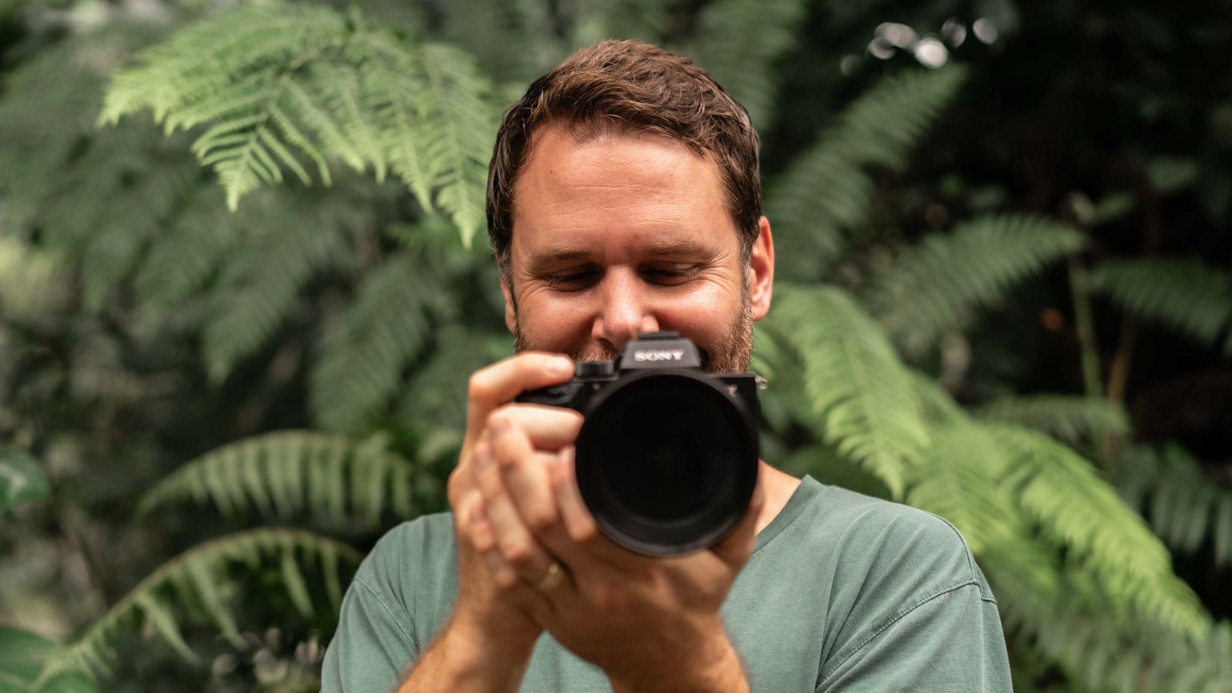 David Lloyd standing outdoors holding a camera, with green foliage in the background | About Page Banner Desktop