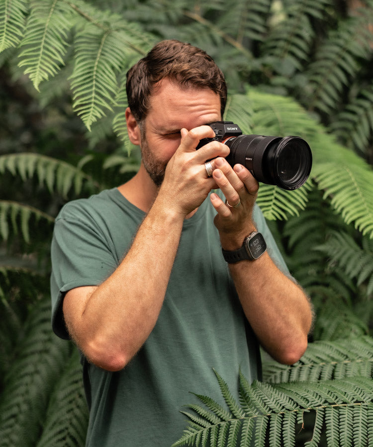 David Lloyd holding a camera to his eye, portrait orientation, greenery background | About Page Banner Mobile