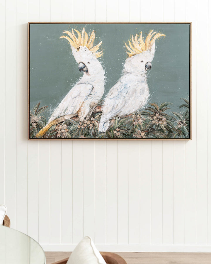 Feathers and Mayhem print of two sulphur-crested cockatoos perched among leafy foliage on a soft grey-green background framed in oak and sitting on a panelled wall above a coastal dining table. Artwork by Stephanie Elizabeth Lloyd of SEA Gallery.