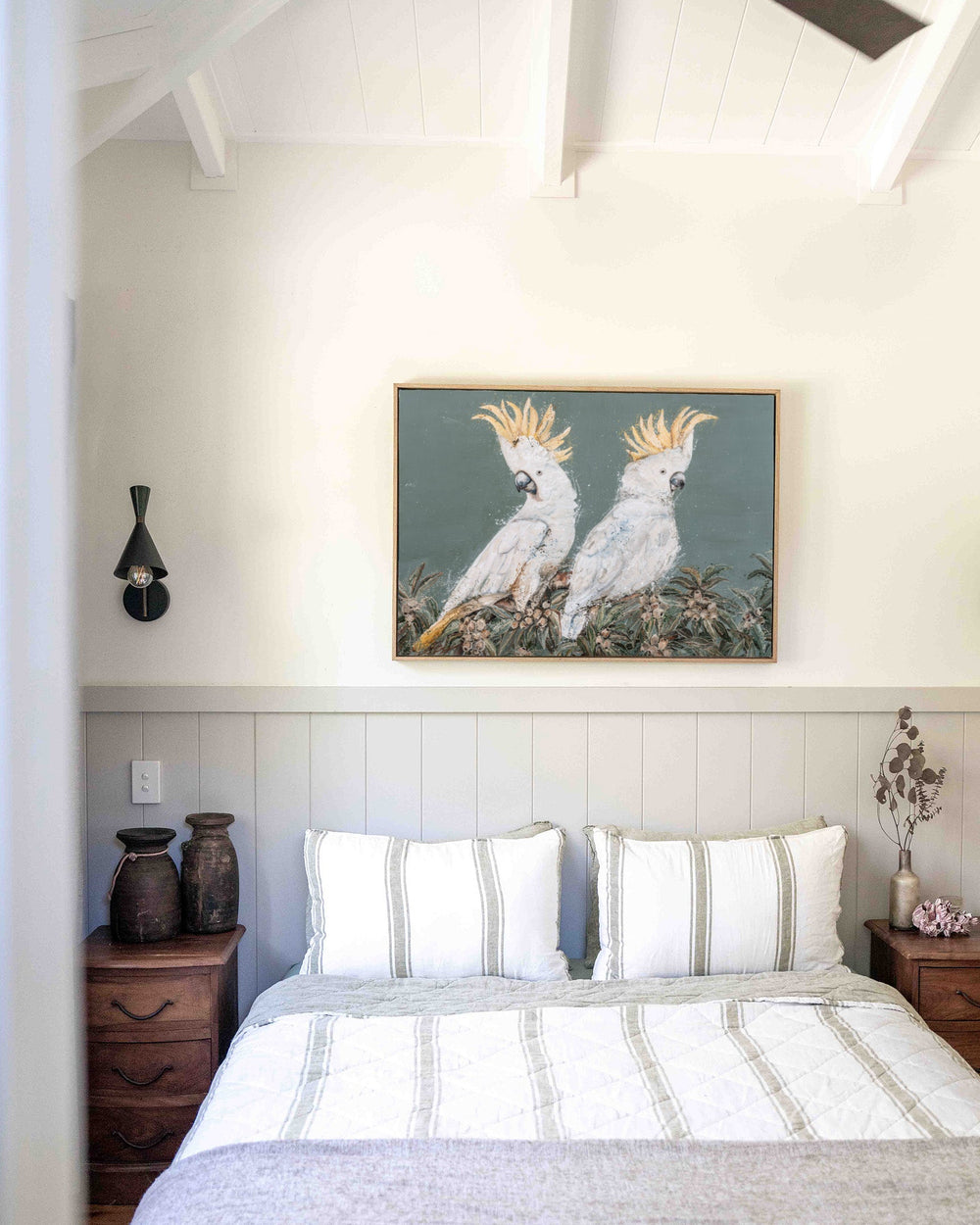 Feathers and Mayhem print of two sulphur-crested cockatoos perched among leafy foliage on a soft grey-green background, in a timber frame above a bed with striped linen.