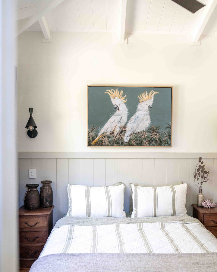 Feathers and Mayhem print of two sulphur-crested cockatoos perched among leafy foliage on a soft grey-green background, in a timber frame above a bed with striped linen.