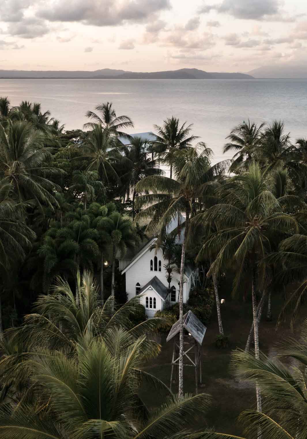 Nestled Amongst The Palms – Aerial photograph of St Mary’s Chapel in Port Douglas surrounded by palms, sunset sky and mountain backdrop | Unframed Rolled Print