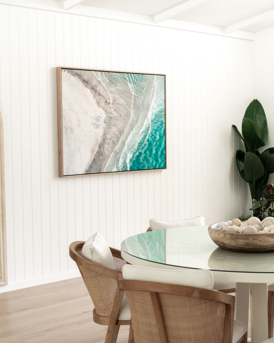 Rattan chairs and a glass table in a coastal dining space, with Pearls and Pelicans artwork on the wall.