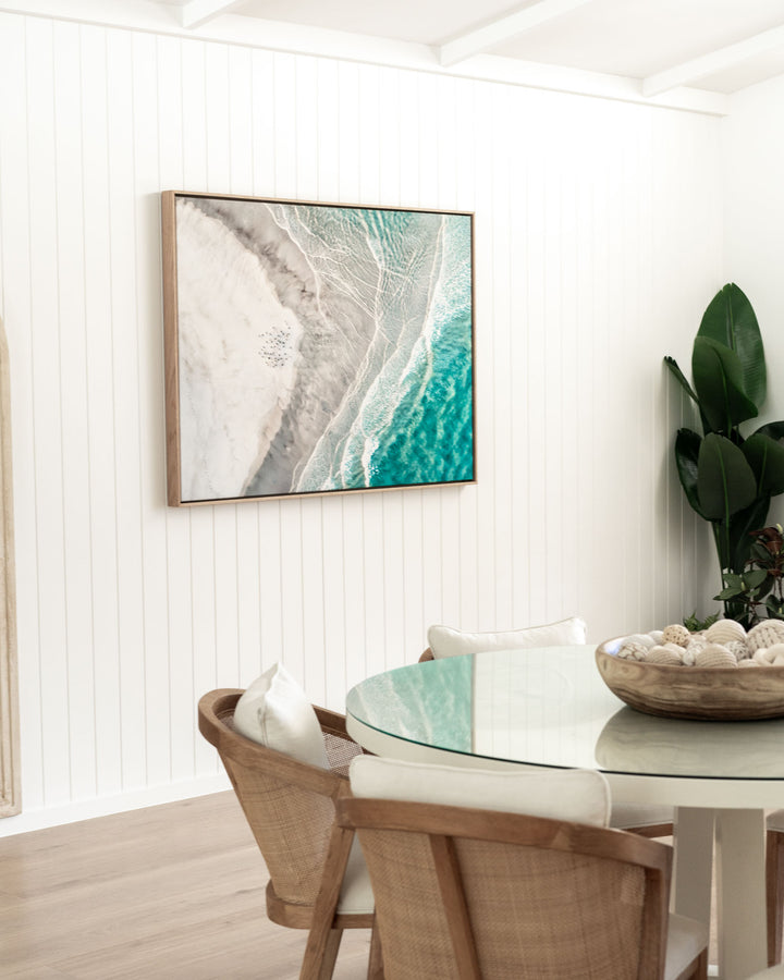 Rattan chairs and a glass table in a coastal dining space, with Pearls and Pelicans artwork on the wall.