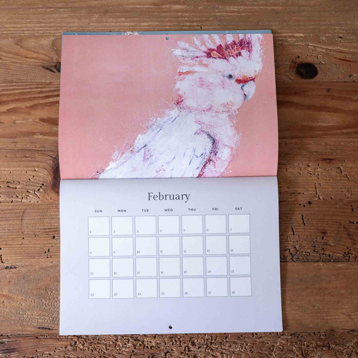 Calendar with a cockatoo illustration on a pink background, placed on a wooden surface.