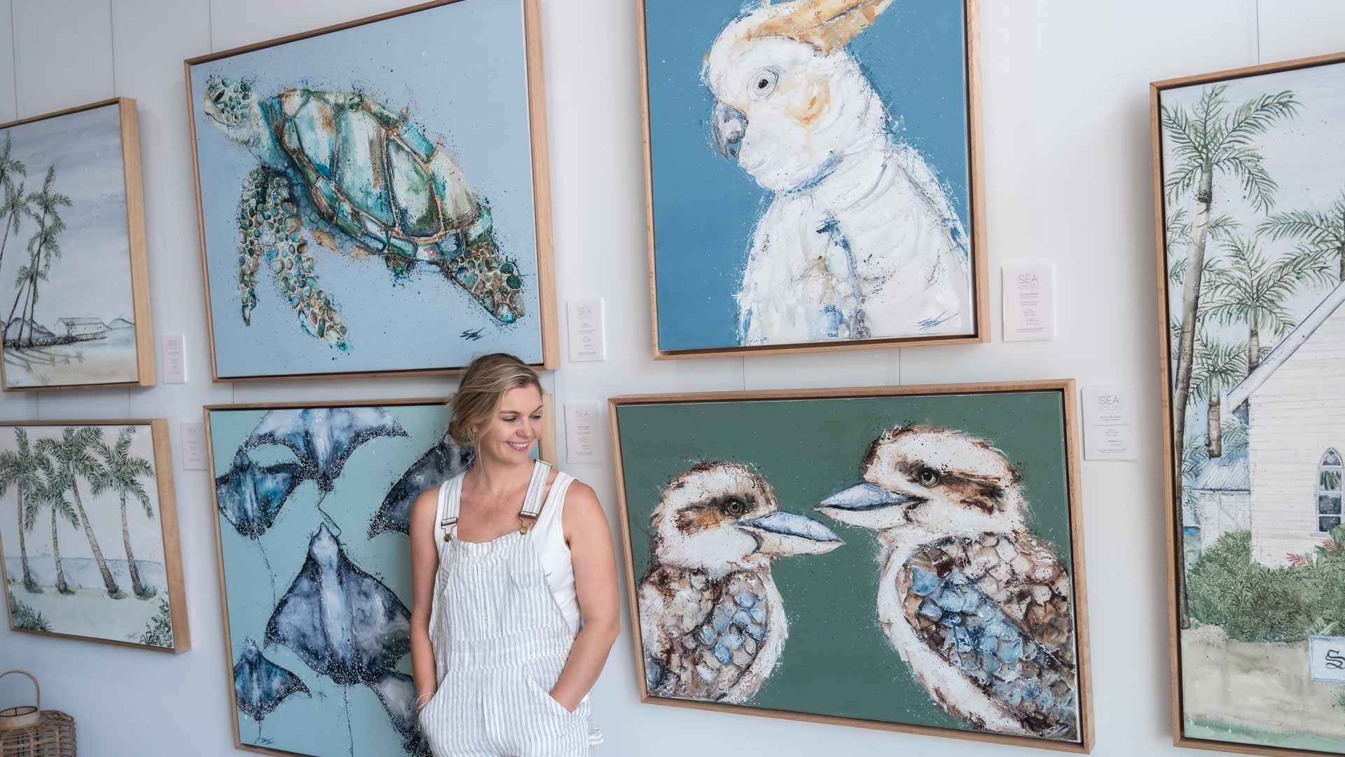 Stephanie Elizabeth – standing next to a wall of her original artworks at SEA Gallery, Port Douglas, showcasing hand-signed one-of-a-kind pieces
