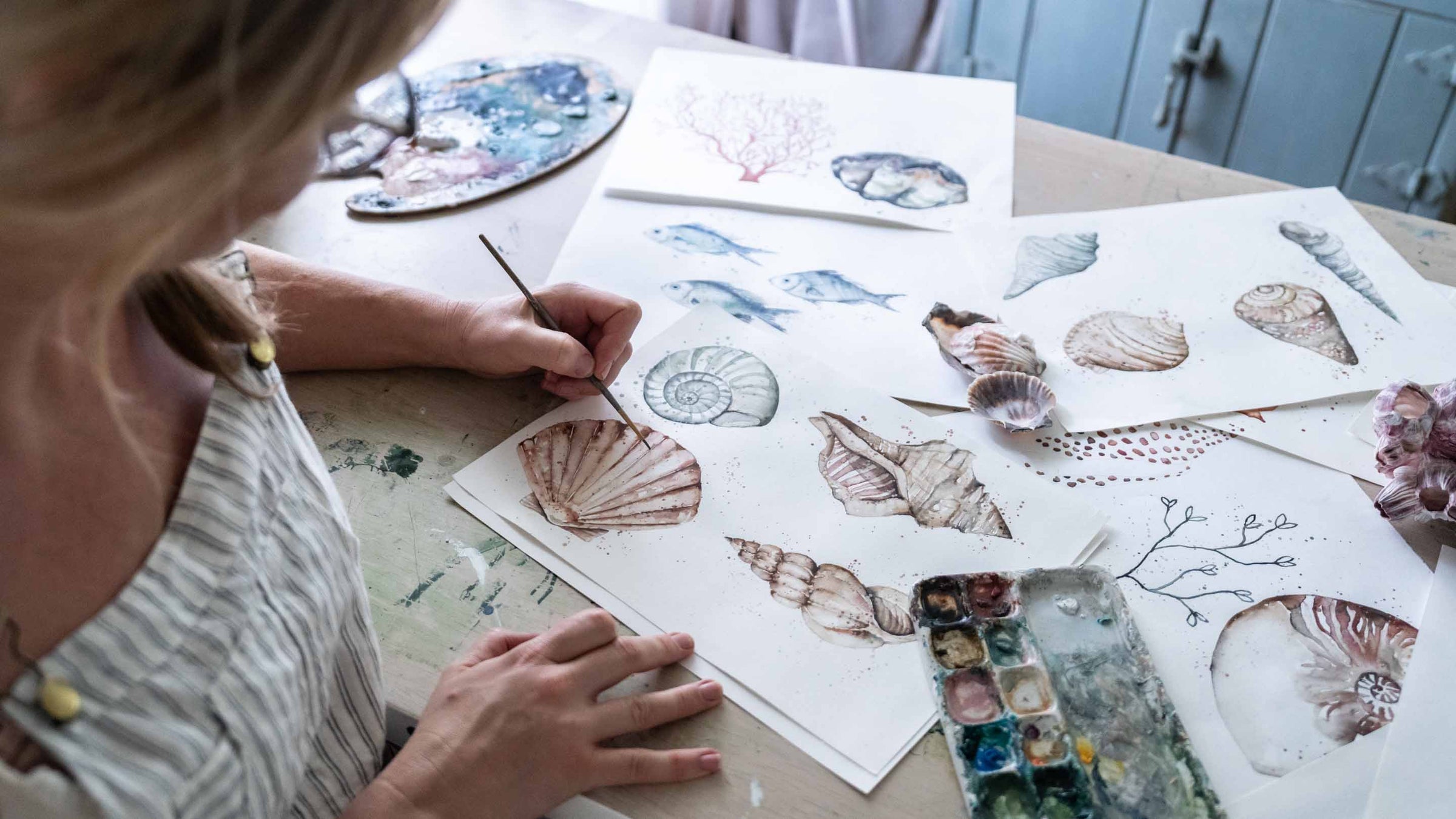 Stephanie Elizabeth painting a collection of sea shells, viewed over her shoulder | About Page Banner Desktop