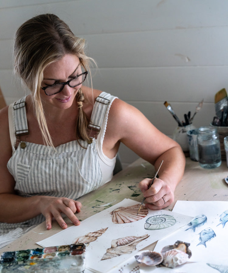 Stephanie Elizabeth smiling while painting a collection of sea shells, facing the camera | About Page Banner Mobile
