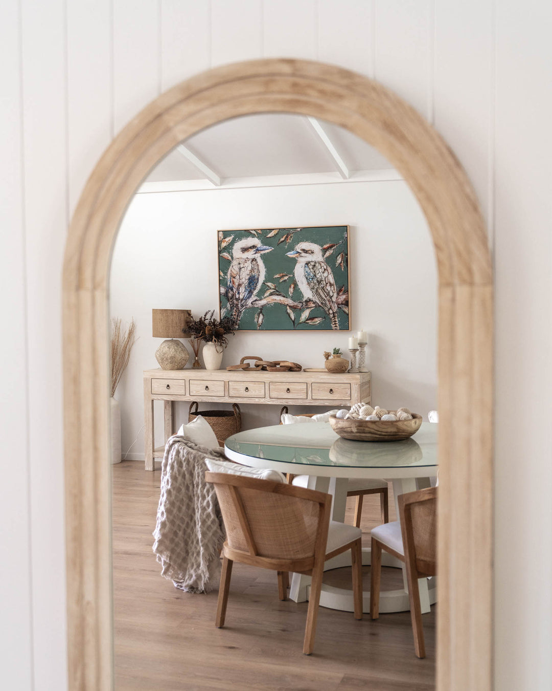 Arched mirror reflecting dining room with wood table and chairs, featuring “We Belong Together | Kookaburras” artwork.